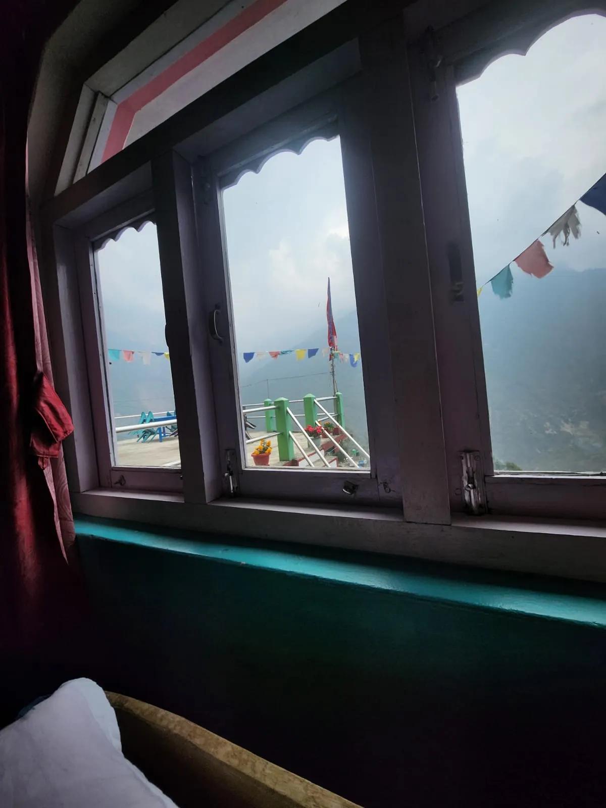 View through a teahouse window in Ulleri showing colourful prayer flags, terrace railing with potted flowers, and misty Himalayan mountains